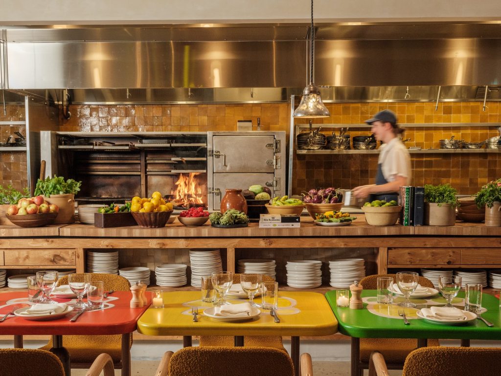 Three colorful tables sit in front of an open restaurant kitchen with chef at work.