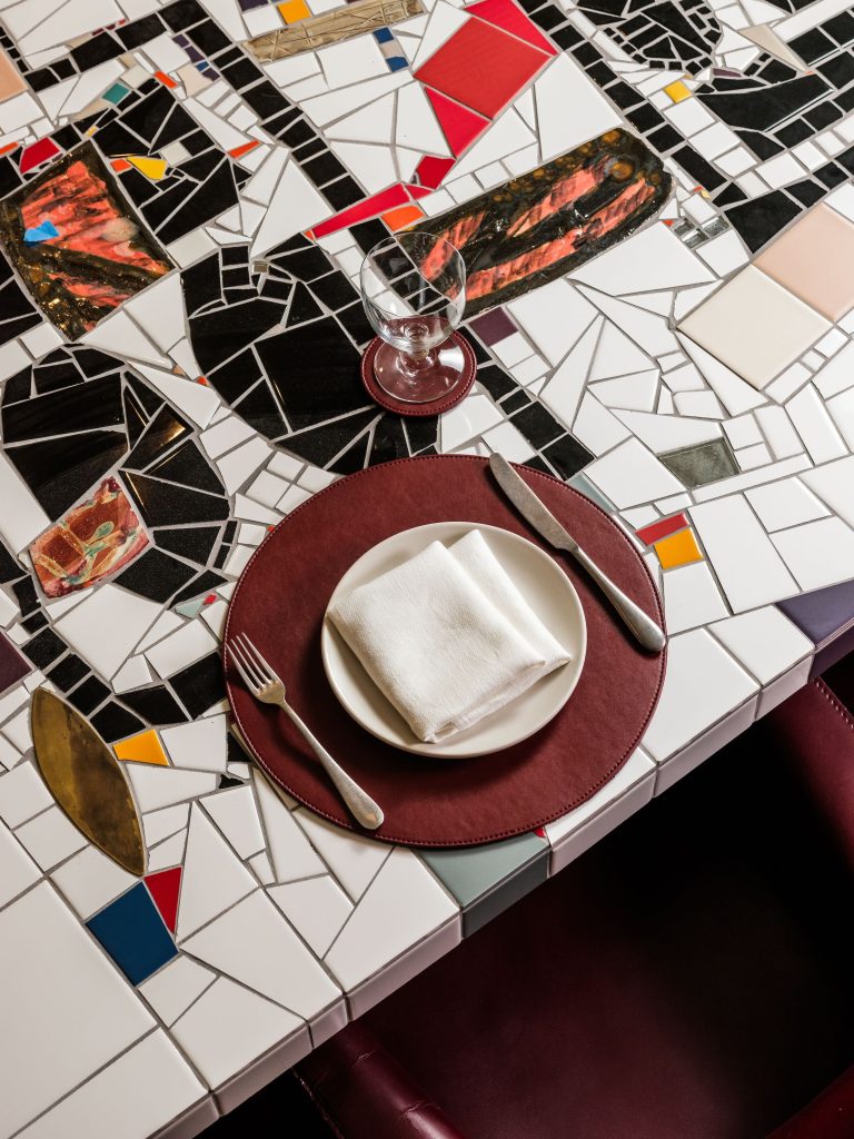 Overhead photo of Private Dining Room Table by Rashid Johnson. The table os a mosaic of multicolored broken tiles that have been pieced back together and there is a table setting on the table.