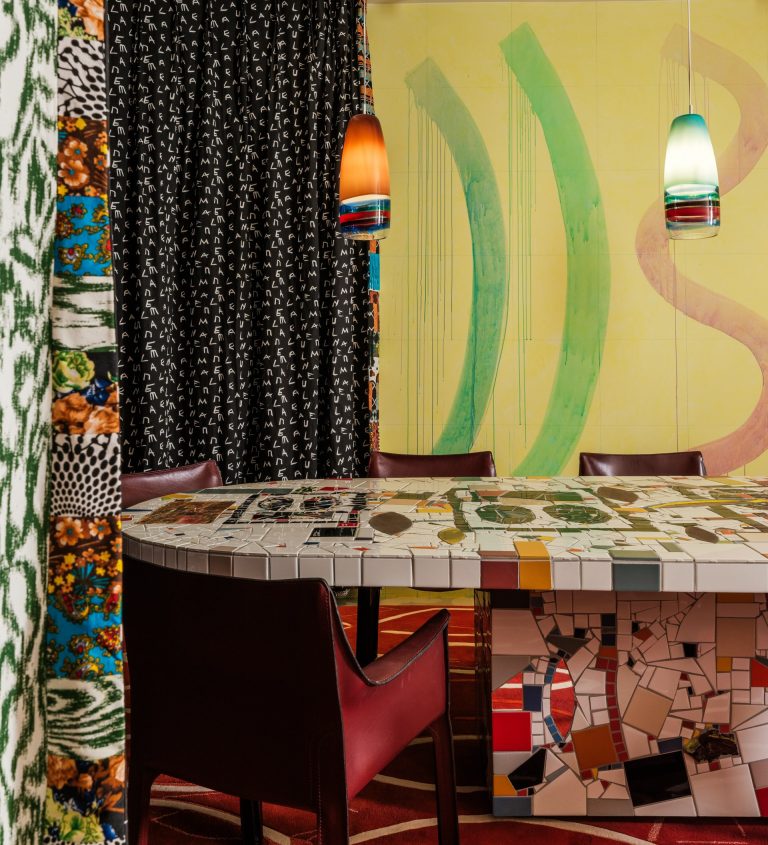 A mosaic table sits in a room with a yellow, green, and pink mural in the back and two light fixtures hanging from the ceiling.