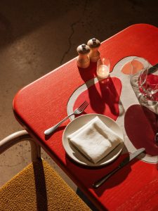 Red table with a white, red, orange and brown design in the center. The table is set for a meal and was created by Mary Heilmann.