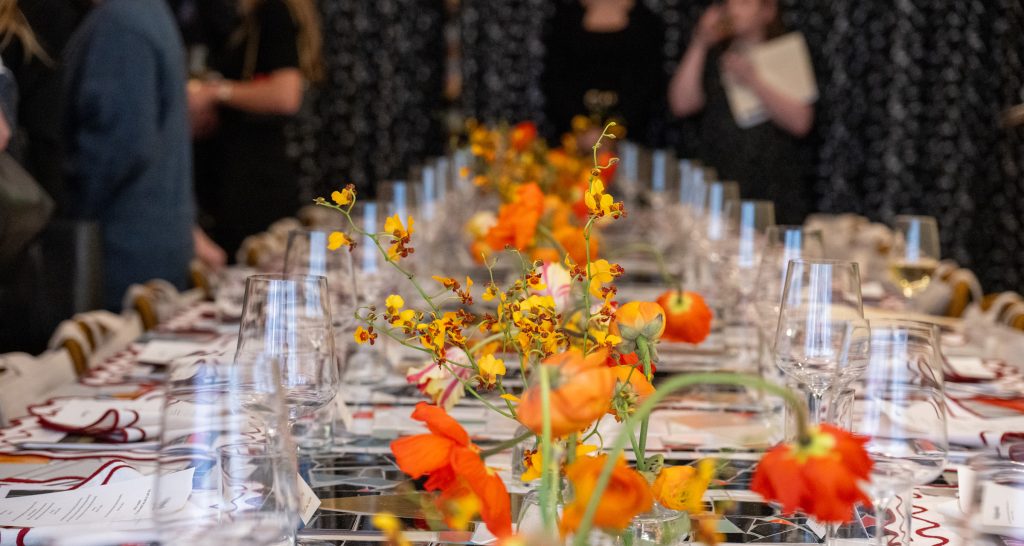 A long table set for dining with red and orange flower arrangements.