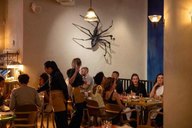 People seated at tables having a meal.The wall behind them is a sculpture of a large spider hanging on the wall.