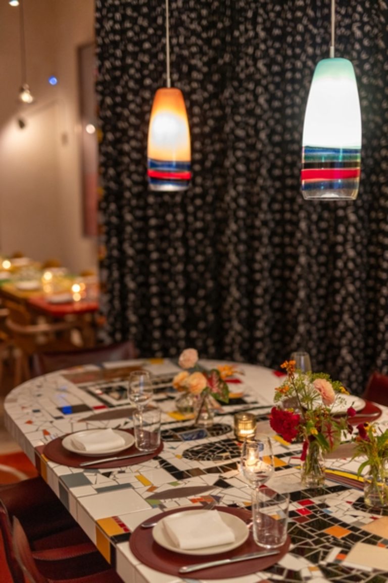 A mosaic table top set for a meal. Two colorful light fixtures hang from the ceiling a black curtain is in the background.