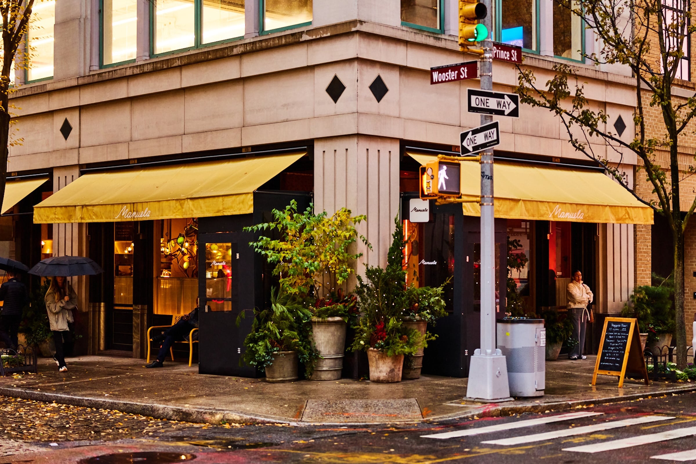 Manuela Exterior on a rainy day. Exterior features yellow awnings and shrubbery on the corner of a city street.