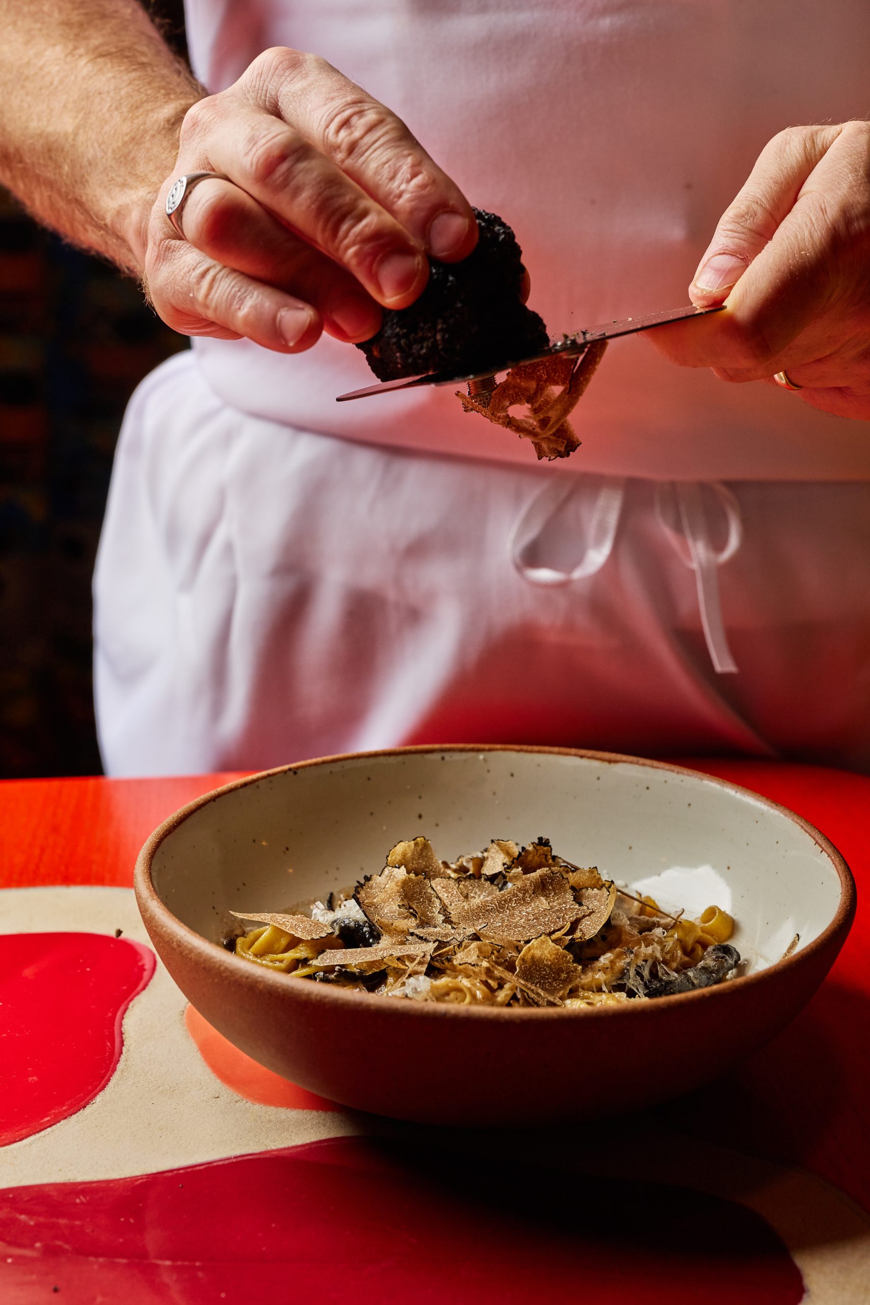 person shaving truffle over pasta bowl on a red table