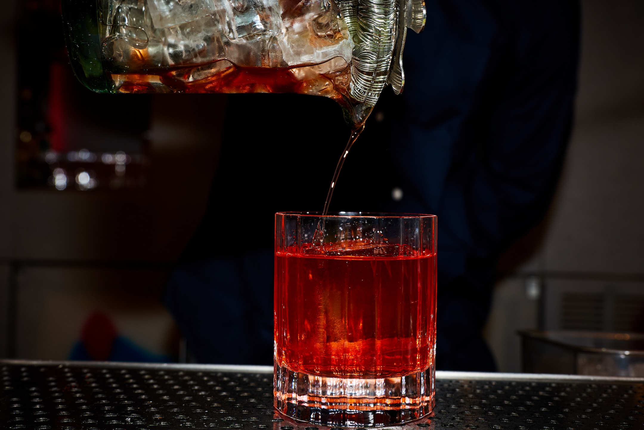 A red cocktail being poured from a mixing glass into a rocks glass.