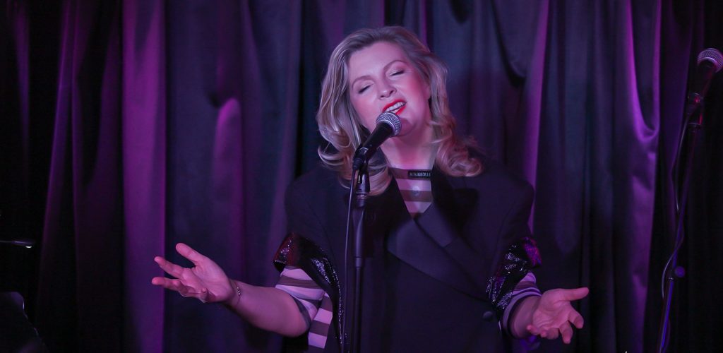 Susanne Mack singing on stage in front of a purple curtain. She is singing into a mic held up by a mic stand.