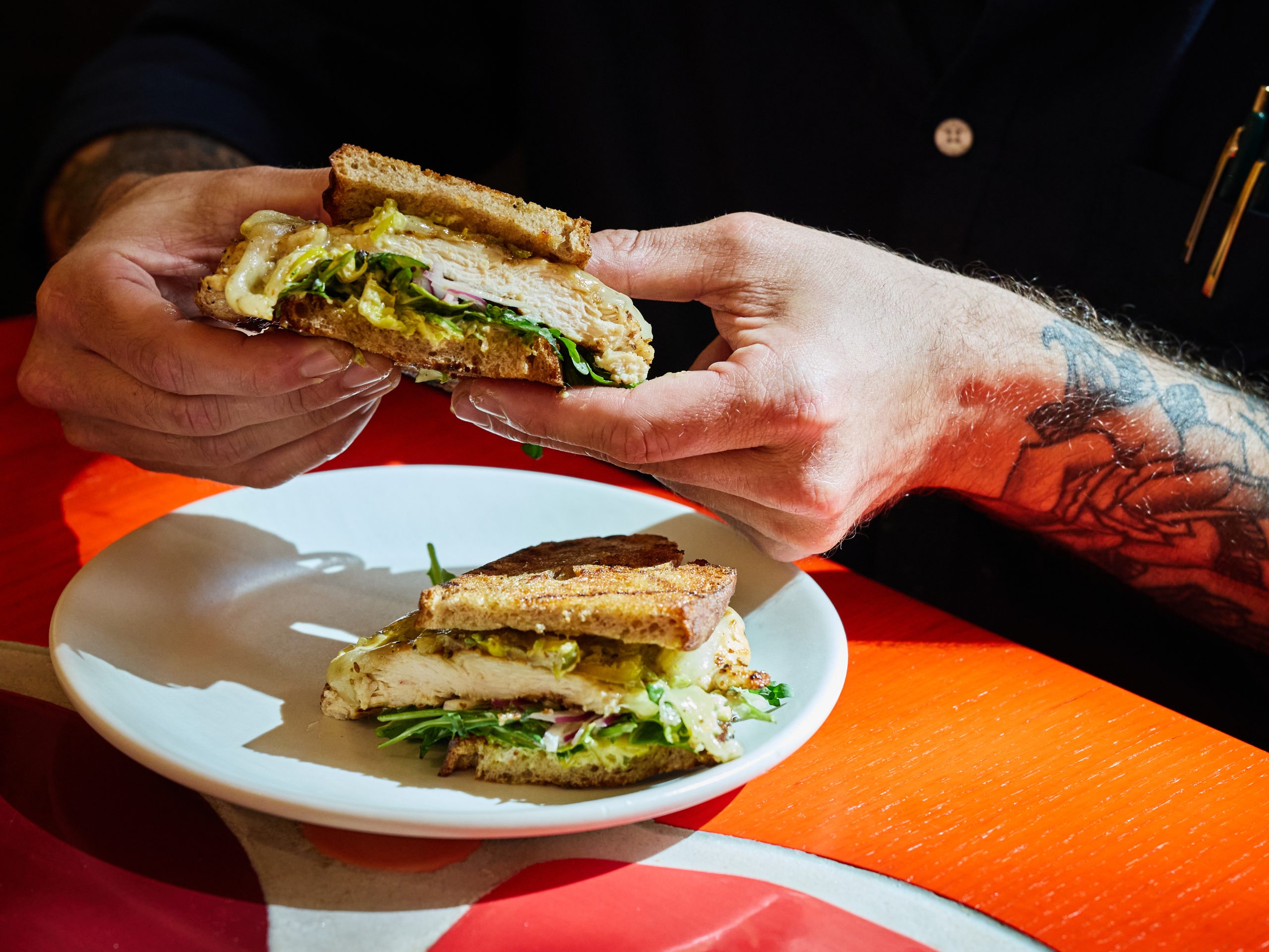 Person holding a buttermilk fried chicken sandwich while seated at a red table inside Manuela restaurant.