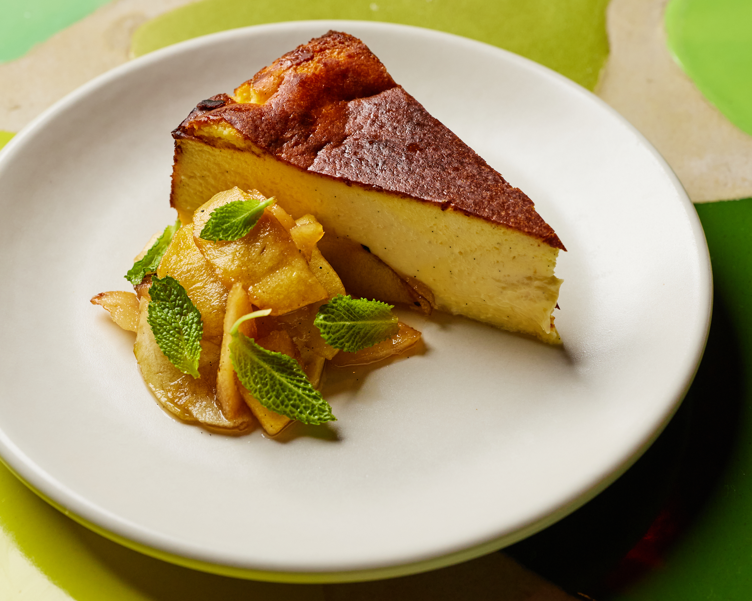 A cheesecake on a white plate. The cheese cake has a brown top and next to is a mixture of yellow fruit and mint.