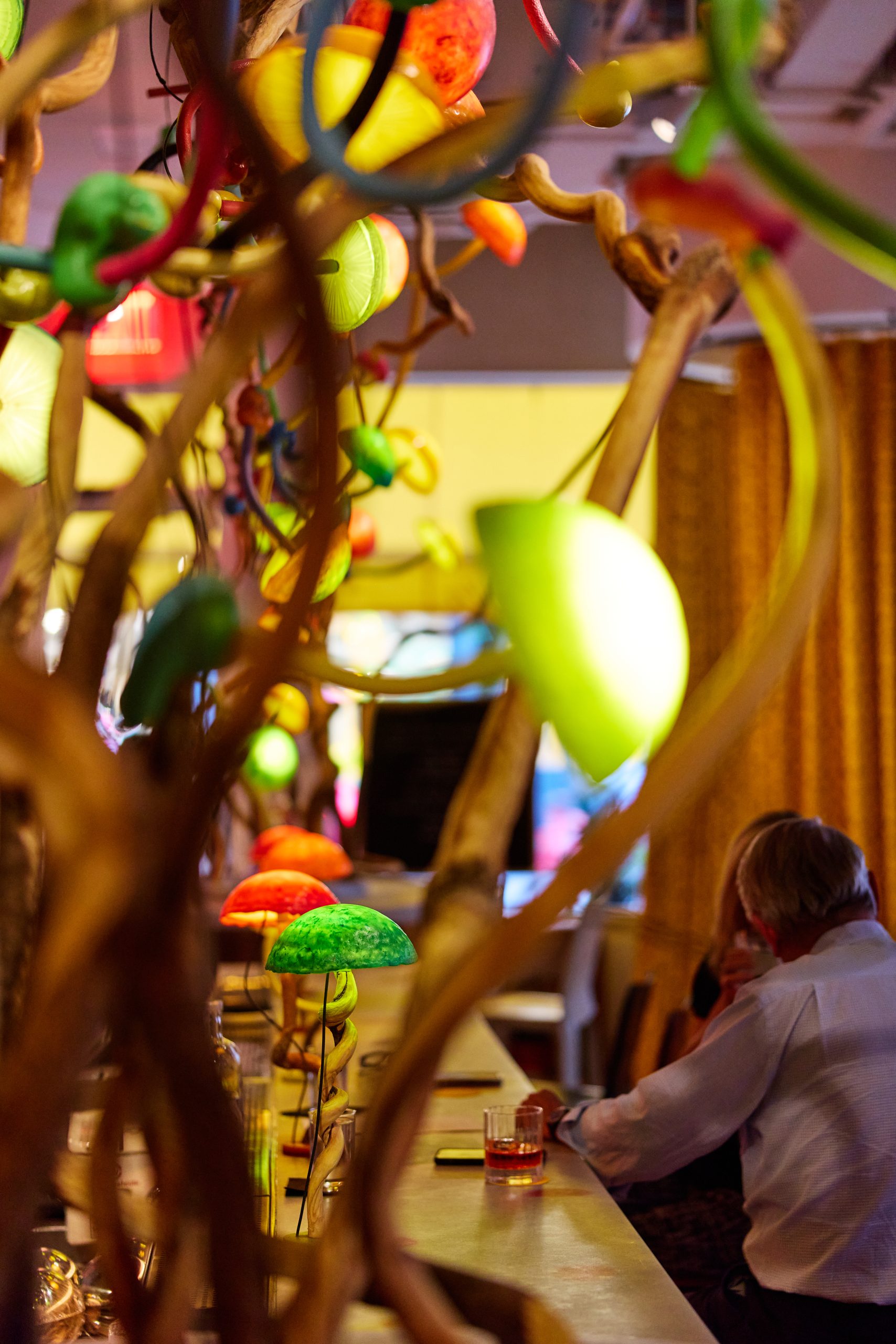 Two people sitting at the bar inside Manuela in SoHo, with a colorful chandelier by artist Mika Rottenberg hanging above them.