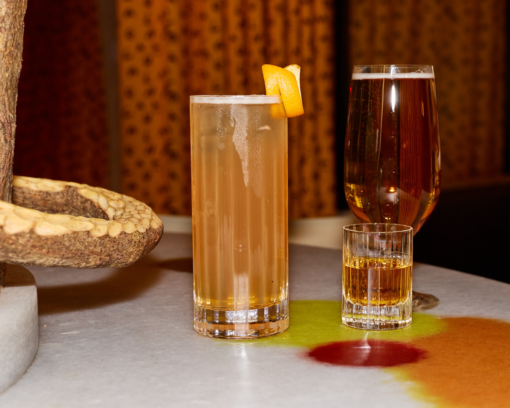 A cocktail in a highball glass sitting next to a glass of beer and a shot glass full of brown whiskey.