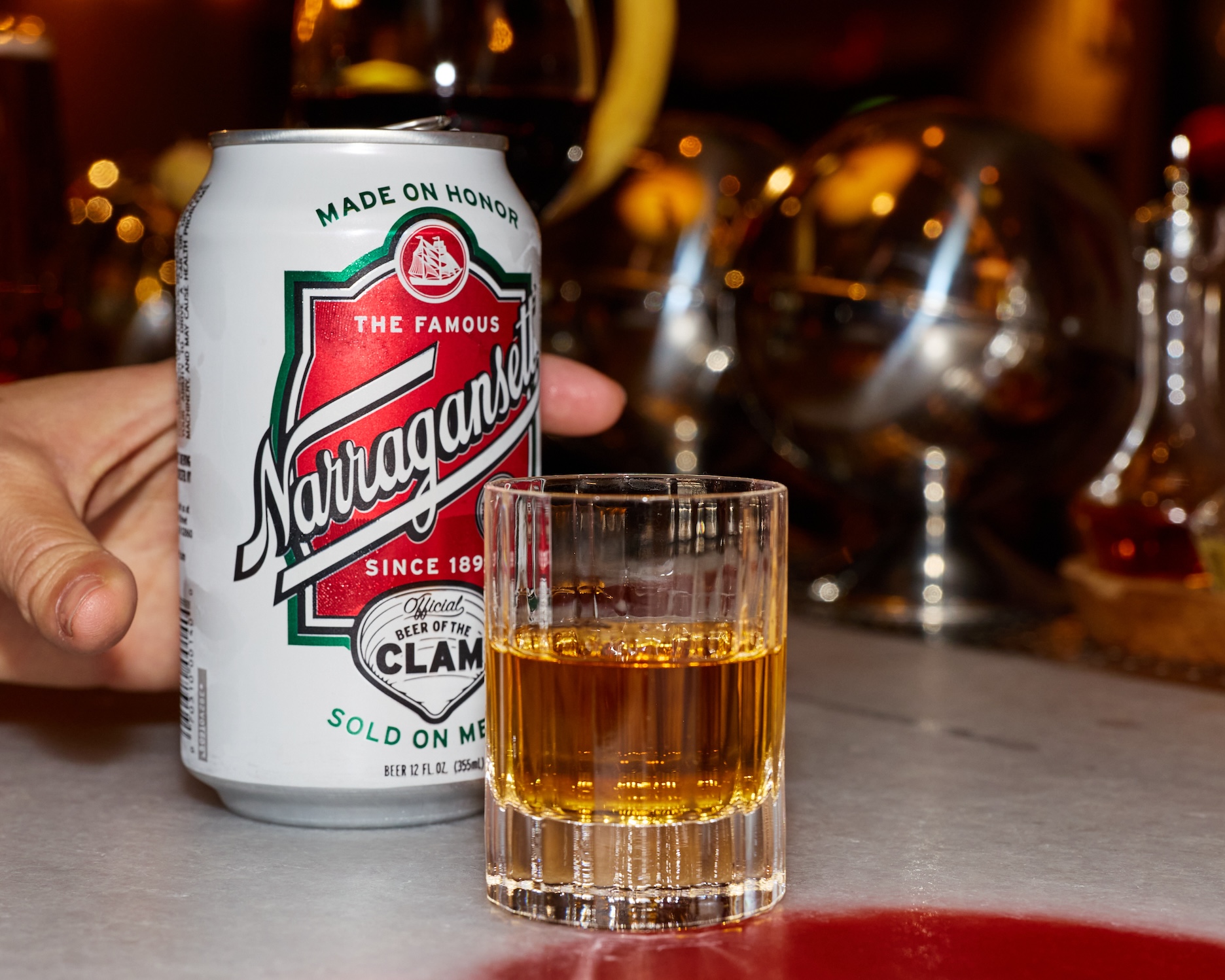 Hand grabbing a can of beer sitting next to a shot glass half full of a brown spirit.