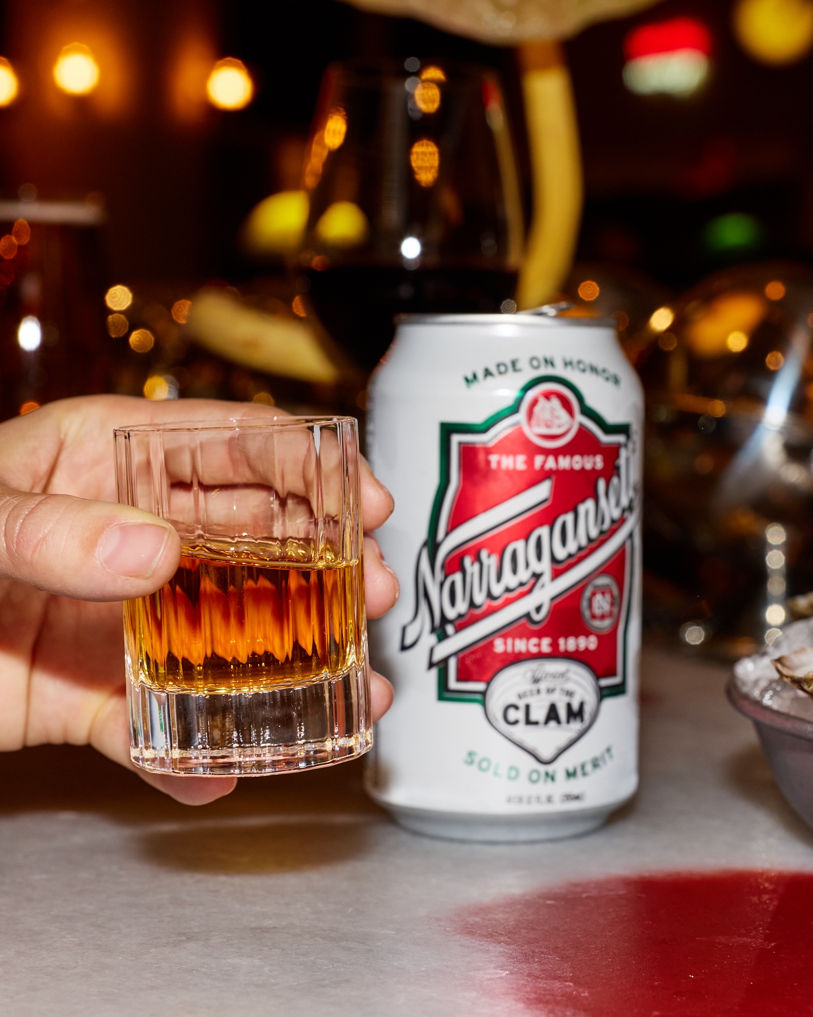 Hand grabbing a shot glass with brown whiskey and a beer can in the foreground.