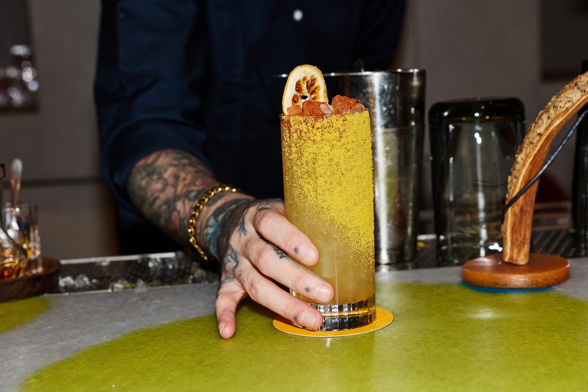 Bar tender placing a tall yellow cocktail in a highball glass on a bar counter, garnished with a dried lemon slice, with a cocktail shaker and glass in the background.
