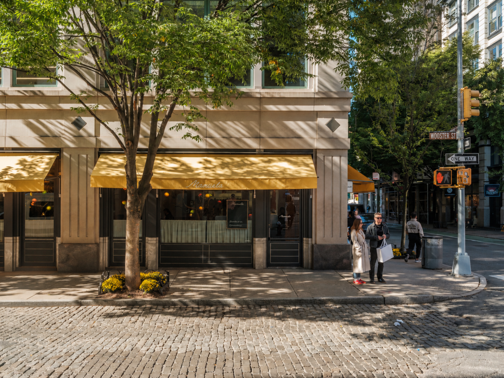 Manuela restaurant with yellow awning on a leafy street corner, cobblestone road in front, and a couple of pedestrians standing near a traffic light and street sign.