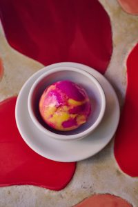 Close-up of a single scoop of yellow and pink ice cream in a small white bowl on a small white plate, sitting on a red patterned table.