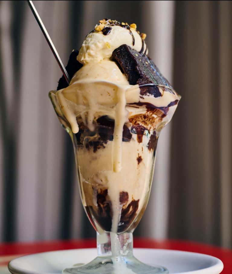 Close-up of brownie & homemade PB ice cream sundae in a tall glass with a metal spoon coming out the top, sitting on a white plate on a red table with beige curtains in the background.