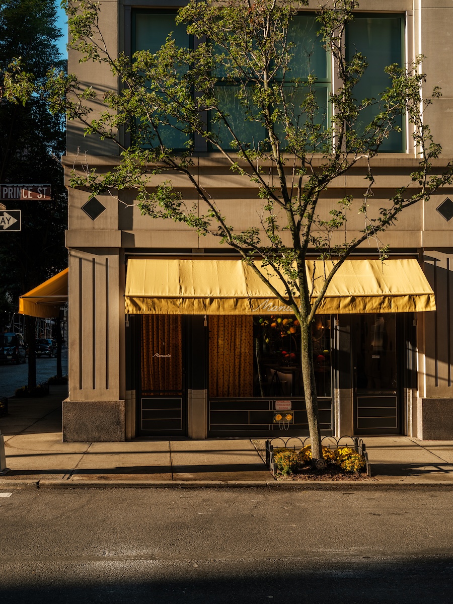 Exterior of Manuela restaurant in SoHo, featuring yellow awnings, large windows, and a tree-lined street frontage.