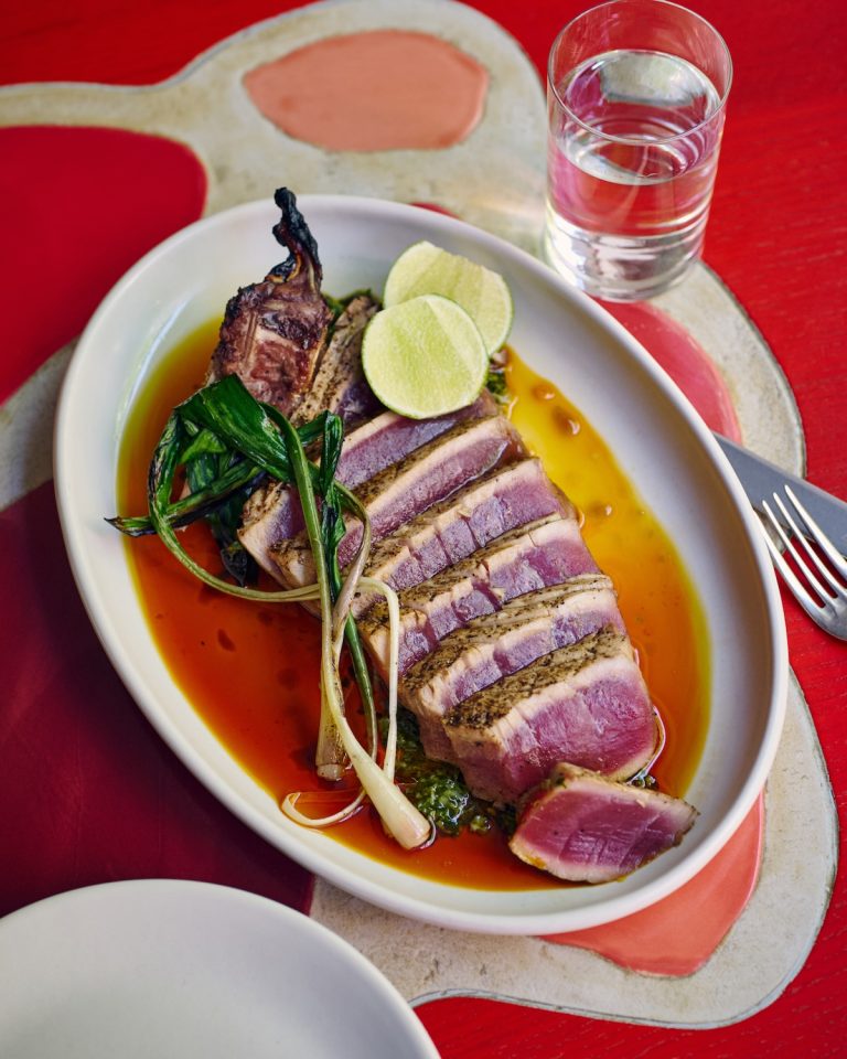 Seared sliced tuna served with vegetables and lime in a shallow white bowl on a red tabletop, next to a glass of water, cutlery and another white plate.
