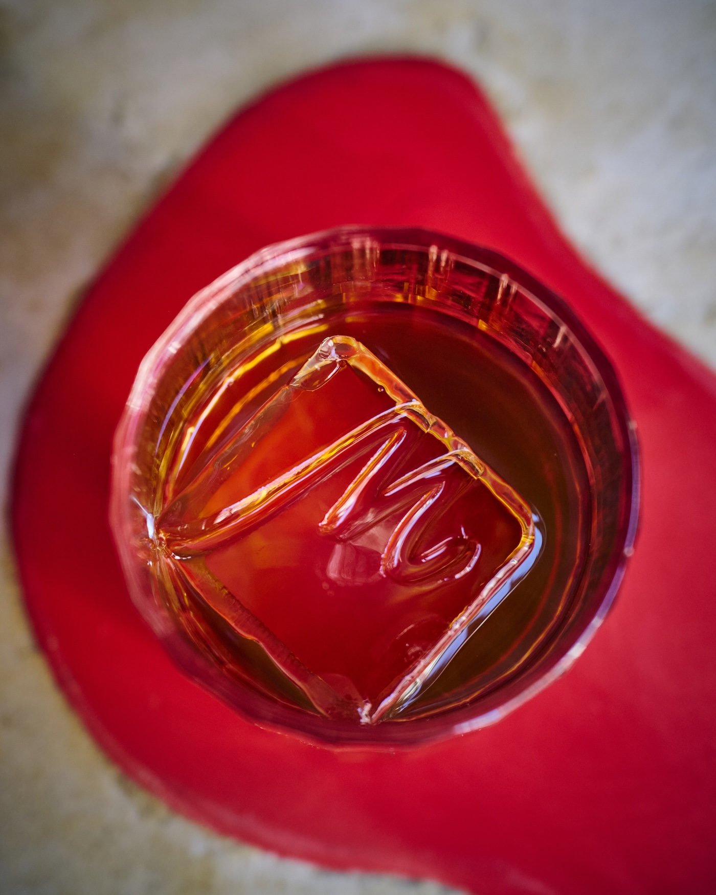 Cocktail served over a large ice cube engraved with an 'M' in a glass, viewed from above on a red tabletop.