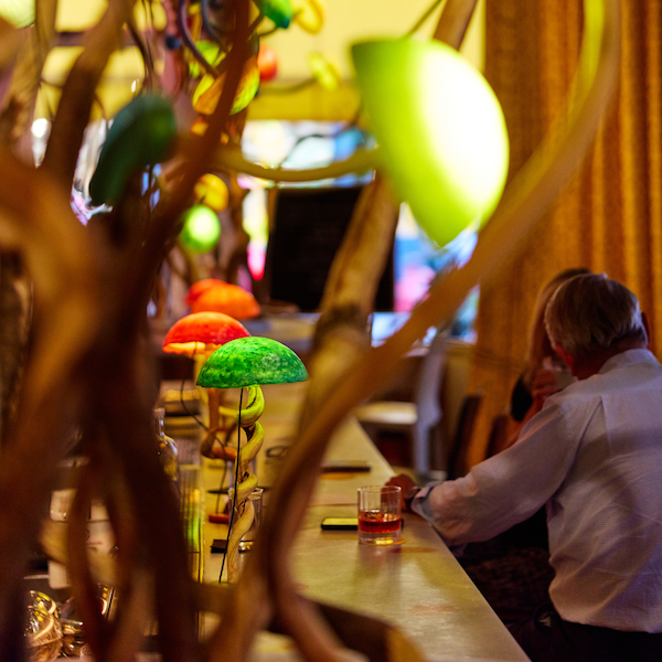 Restaurant interior with twisting branch structures framing a long counter lined with colorful illuminated lamps, while guests sit and drink in the background before dinner.