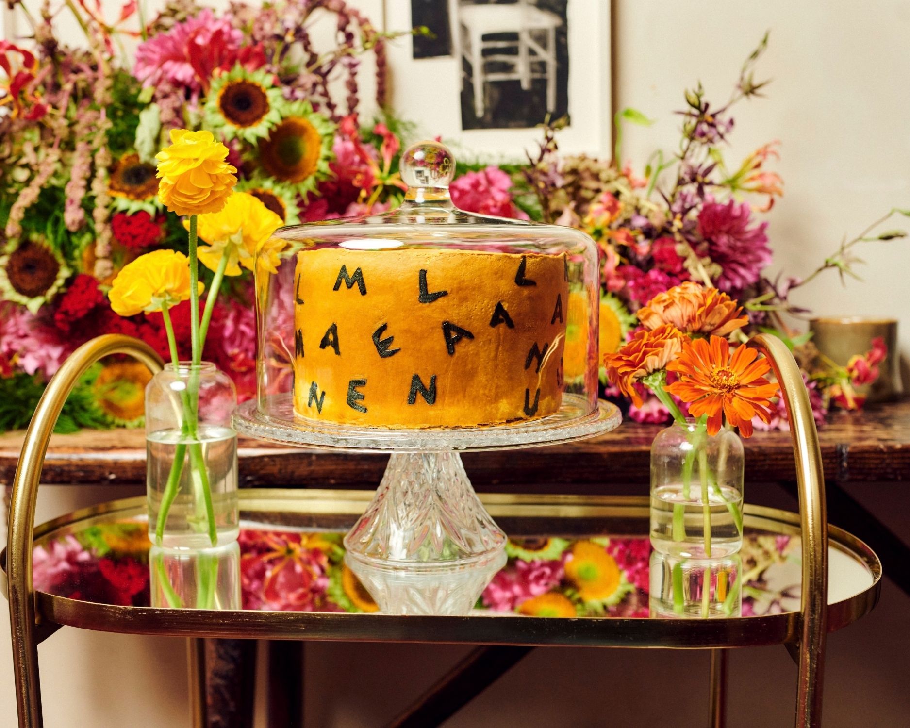 Golden cake with scattered black letters under a glass cloche, surrounded by bright flowers on a brass bar cart.