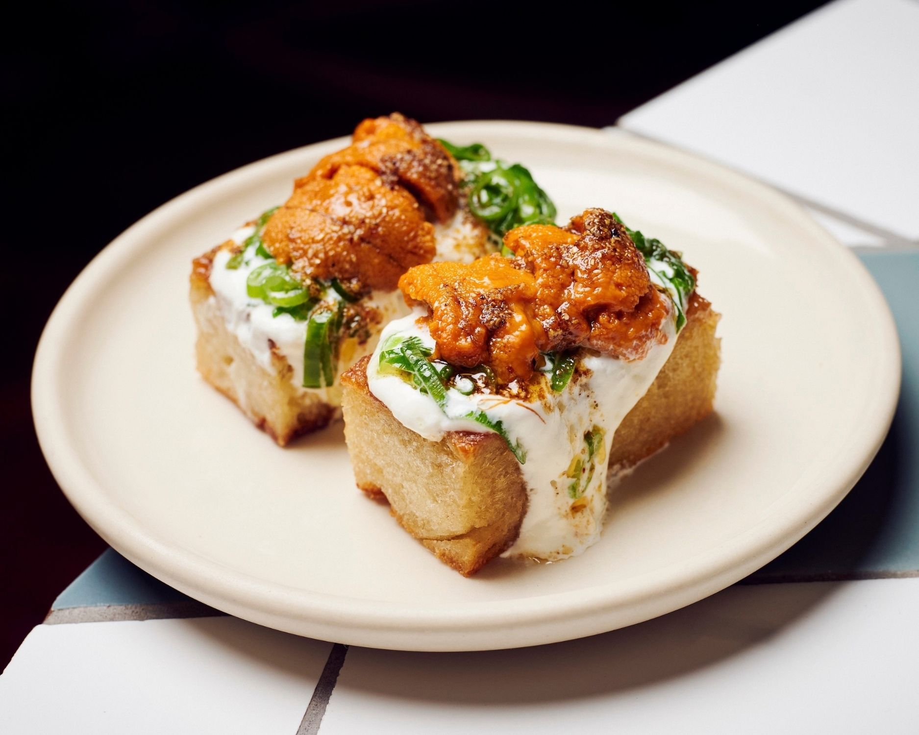 Two toasted bread squares topped with creamy white spread, sliced green chilies, and fresh sea urchin on a cream plate.